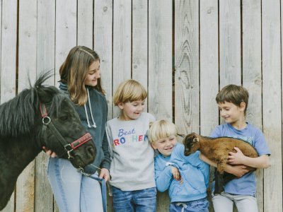 Vier Kinder vor einem Holzzaun, eines hält ein kleines braunes Zicklein, ein anderes führt ein schwarzes Pony an der Leine.