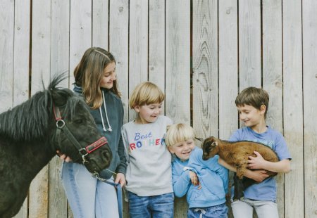 Vier Kinder vor einem Holzzaun, eines hält ein kleines braunes Zicklein, ein anderes führt ein schwarzes Pony an der Leine.