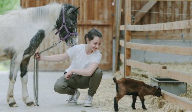 Mädchen hockt neben Pony und sieht Zicklein an, das an Heu in einem Stall frisst