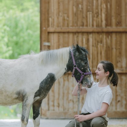 Ein Mädchen kniet vor einem gescheckten Pony und hält dessen Führstrick in einer Scheune.