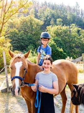 Kind mit Helm sitzt auf braunem Pferd, Mädchen hält Pferd an der Leine, Wald im Hintergrund