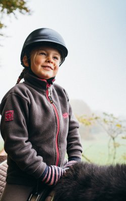 Kind mit Reithelm und Handschuhen sitzt auf einem Pferd im Freien an einem nebligen Tag