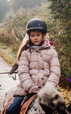 Mädchen mit Helm reitet Pony auf einem Weg, weitere Kinder und Ponys im Hintergrund