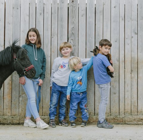 Vier Kinder stehen vor einem Holzzaun, eines hält ein kleines Pony, ein anderes ein Zicklein