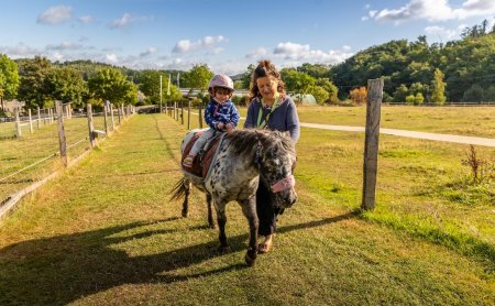 Kind mit Helm reitet Pony auf einem grasbewachsenen Weg, Frau führt das Pony an der Seite