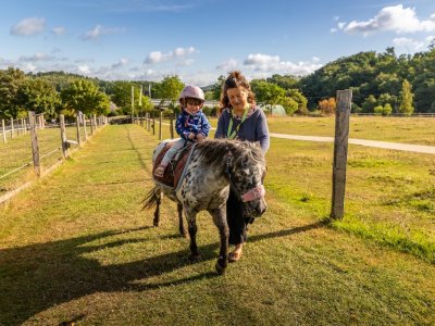 Kind mit Helm reitet Pony auf einem grasbewachsenen Weg, Frau führt das Pony an der Seite