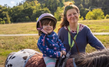 Kind mit Helm auf Pony sitzend, lächelnde Frau steht daneben auf einer Wiese