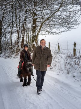 Mann führt Pony mit Kind im Helm auf verschneitem Weg neben Bäumen und Zaun