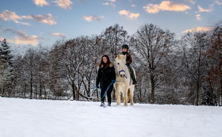 Frau führt Kind auf weißem Pferd durch verschneite Landschaft bei Sonnenuntergang