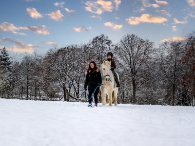 Frau führt Kind auf weißem Pferd durch verschneite Landschaft bei Sonnenuntergang
