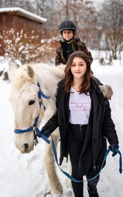 Mädchen führt weißes Pferd im Schnee, Junge mit Helm sitzt auf dem Pferd im Hintergrund