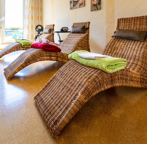 Drei geflochtene Liegen mit bunten Handtüchern in einem hellen Raum mit Pflanzen und Bildern an der Wand