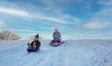 Kinder rodeln nebeneinander den verschneiten Hang unter blauem Himmel hinunter