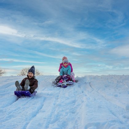 Kinder rodeln nebeneinander den verschneiten Hang unter blauem Himmel hinunter
