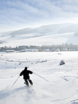 Skifahrer fährt einen schneebedeckten Hang hinunter mit Dorf und Bergen im Hintergrund