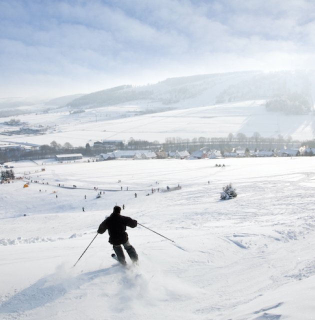 Skifahrer fährt einen schneebedeckten Hang hinunter mit Dorf und Bergen im Hintergrund