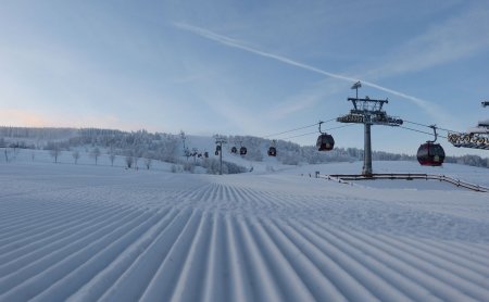 Gepflegte Skipiste mit Gondelbahn rechts und verschneiten Bäumen im Hintergrund unter blauem Himmel
