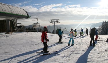 Skifahrer stehen auf schneebedecktem Hang bei Sessellift unter sonnigem Himmel mit Bergen im Hintergrund