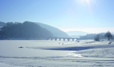 Verschneite Landschaft mit gefrorenem See, Brücke im Hintergrund und strahlender Sonne am Himmel