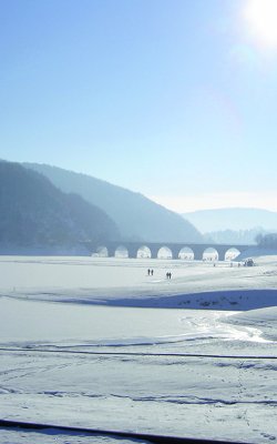 Verschneite Landschaft mit gefrorenem See, Brücke im Hintergrund und strahlender Sonne am Himmel