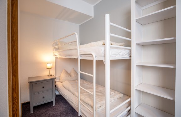 Weißes Etagenbett mit Bettwäsche neben grauem Nachttisch mit Lampe und leerem Regal