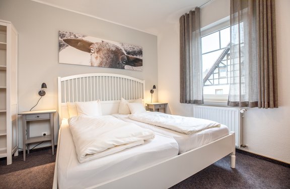 Helles Schlafzimmer mit weißem Doppelbett, zwei Nachttischen, Fenster mit Vorhängen und Bild einer Kuh über dem Bett