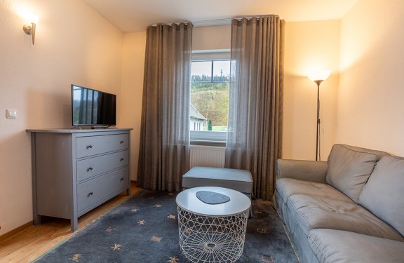 Wohnzimmer mit grauem Sofa rechts, rundem Tisch in der Mitte, grauem Sideboard links und Fenster mit Vorhängen