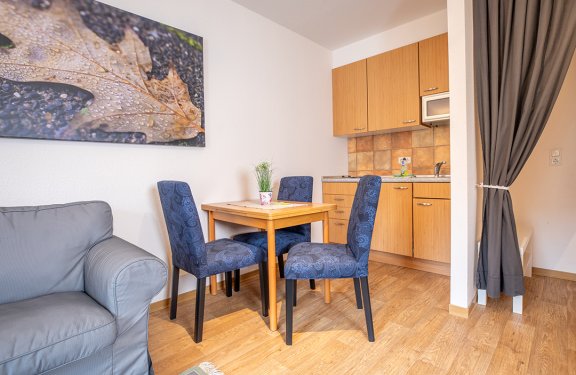 Essbereich mit Holztisch und blauen Stühlen neben einer kleinen Küche mit Holzschränken