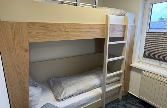 Hochbett mit Leiter neben Fenster und blauem Hocker auf blauem Teppichboden in kleinem Zimmer