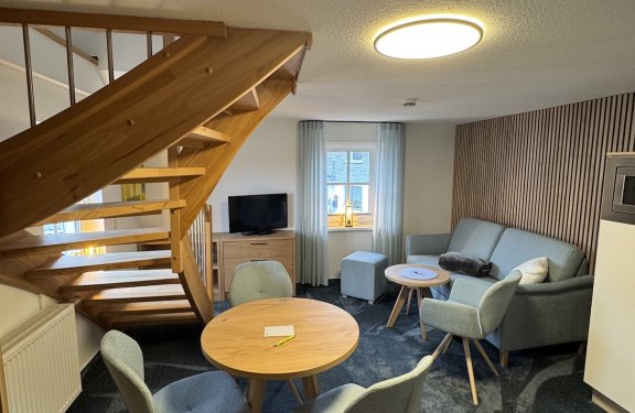 Wohnzimmer mit Holztreppe links, runden Tischen und grauen Stühlen, Sofa rechts vor Fenster