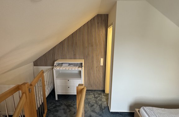 Schlafzimmer mit Dachschräge, Treppengeländer links, Wickelkommode und Babybett an der Wand