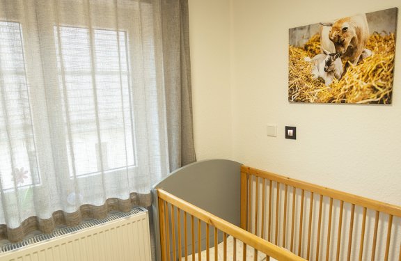 Kinderzimmer mit Holzgitterbett, grauen Vorhängen, grünem Hocker und Bild mit Ziegen an der Wand