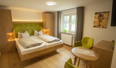 Helles Hotelzimmer mit Doppelbett, grüner Polsterung, zwei grünen Sesseln und Fenster mit Vorhängen