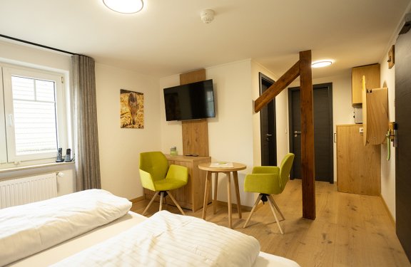 Helles Hotelzimmer mit zwei Betten, zwei grünen Stühlen, kleinem Holztisch und Wandfernseher
