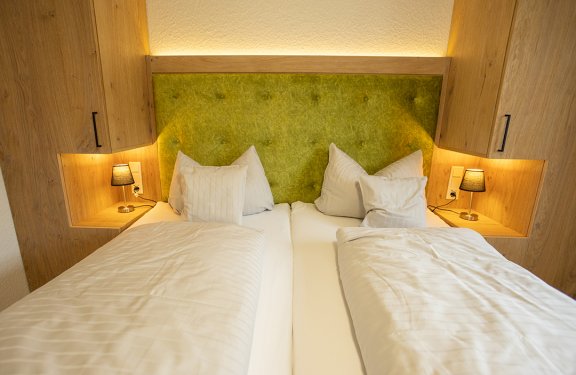 Zwei Einzelbetten mit weißen Bettdecken und Kissen vor grünem gepolstertem Kopfteil, Holzschränke und Nachttischlampen