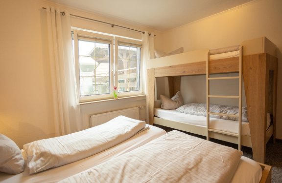 Helles Schlafzimmer mit Etagenbett rechts und Einzelbett links vor Fenster mit weißen Vorhängen