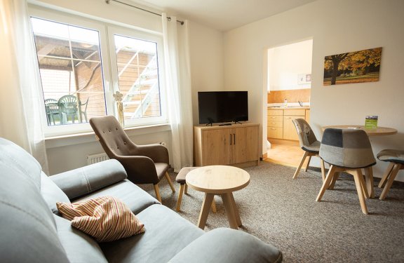 Helles Wohnzimmer mit Sofa, Sessel, kleinem Holztisch, TV auf Sideboard und Blick in die Küche
