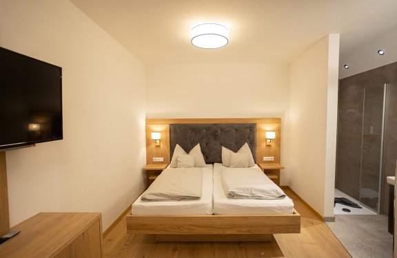 Hell eingerichtetes Schlafzimmer mit Doppelbett, zwei Nachttischen, Wandlampen und Fernseher an der Wand