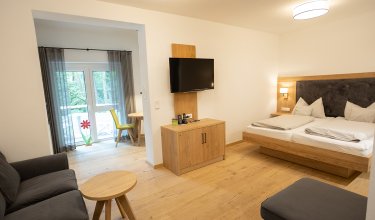 Helles Hotelzimmer mit Doppelbett rechts, Fernseher an der Wand und Sitzecke links vor Fenster mit Vorhängen