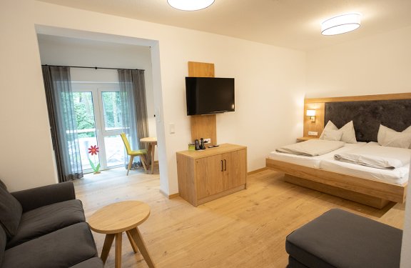 Helles Hotelzimmer mit Doppelbett rechts, Fernseher an der Wand und Sitzecke links vor Fenster mit Vorhängen