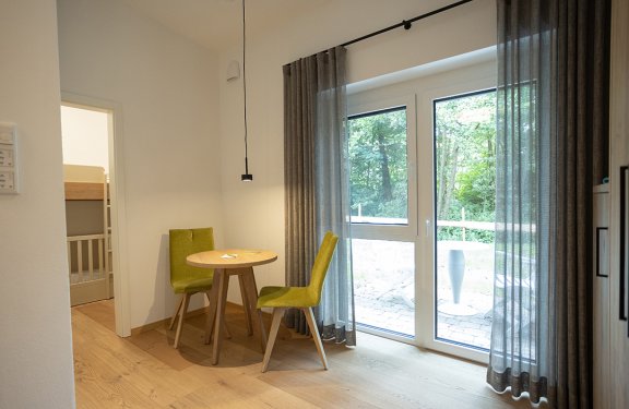 Kleiner runder Holztisch mit zwei grünen Stühlen neben einer großen Glas-Schiebetür mit grauen Vorhängen