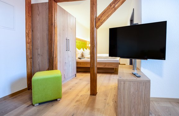 Blick in ein Zimmer mit grünem Hocker links, Holzschrank und Bett im Hintergrund, rechts ein Fernseher auf Holzkommode