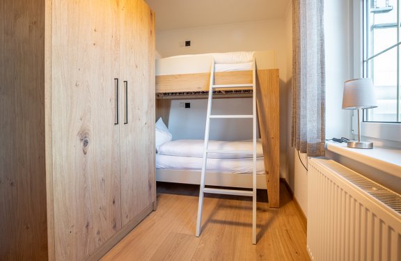 Helles Schlafzimmer mit Etagenbett, Holzschrank links und Fenster mit Lampe rechts