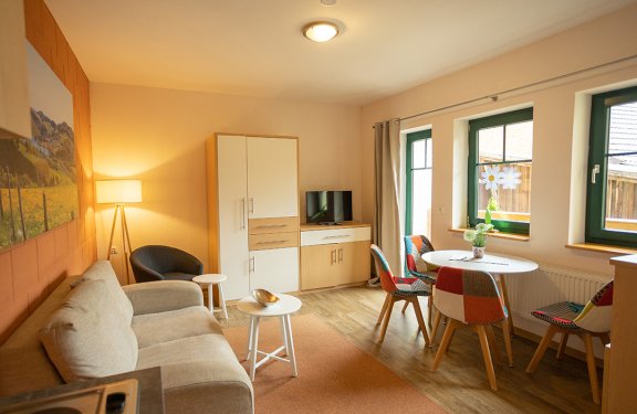 Helles Wohnzimmer mit Sofa links, Stehlampe, Schrank und Fernseher in der Mitte, bunter Essbereich rechts vor Fenstern