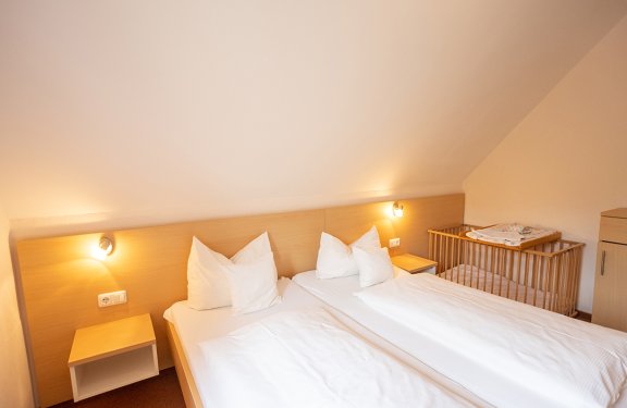 Zwei Einzelbetten mit weißen Kissen und Decken unter einer schrägen Decke, Babybett rechts