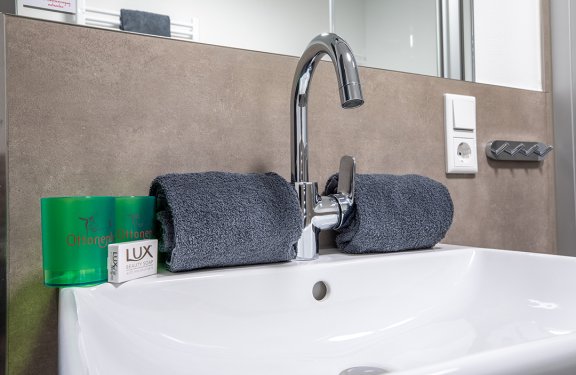 Badezimmerwaschbecken mit Chromarmatur, zwei grauen Handtüchern, grünen Bechern und Lux Beauty Soap