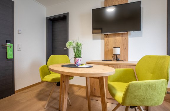 Moderner Raum mit rundem Holztisch, zwei grünen Stühlen, TV an der Wand und Tür mit grünem Schild