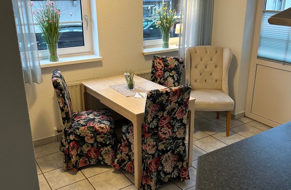 Essbereich mit quadratischem Holztisch, drei Stühlen mit Blumenmuster und einem beigen Sessel vor zwei Fenstern mit Blumenvasen