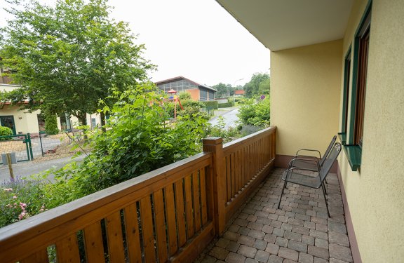 Balkon mit zwei Metallstühlen rechts, Holzzaun links und grüner Garten mit Bäumen und Straße im Hintergrund