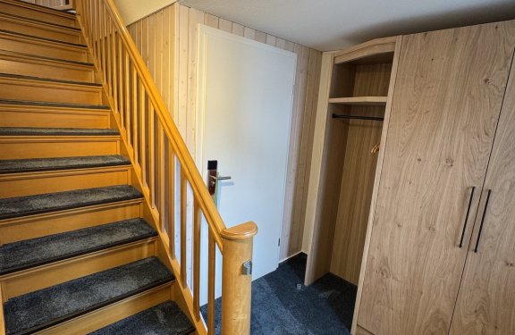 Holztreppe mit dunkelgrauem Teppich links, weiße Tür und Holzkleiderschrank rechts in Flur